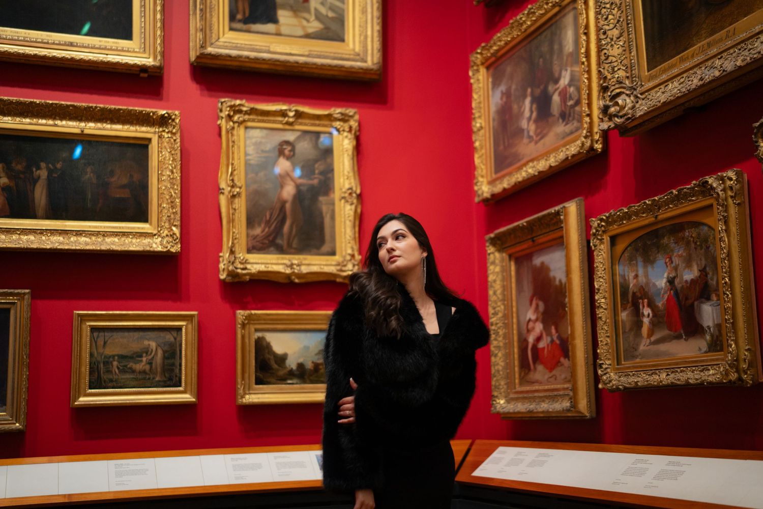 A person in black contemplates artwork in a gallery with red walls and ornate gold frames.