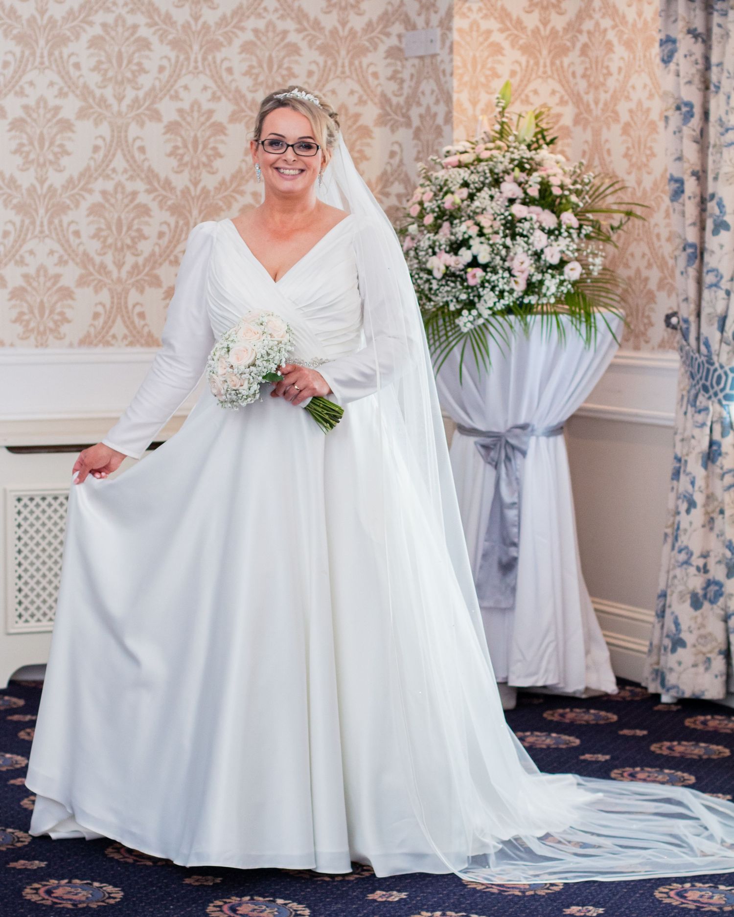 Bride in elegant white wedding gown posing in venue with ornate wallpaper backdrop.