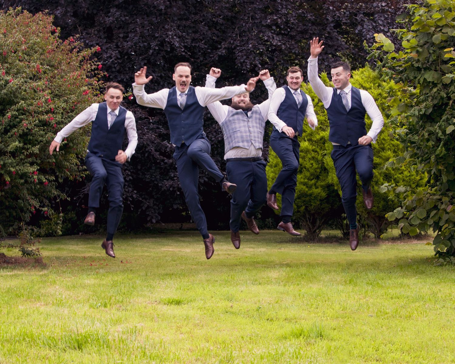 Groomsmen jumping in unison on grass during outdoor wedding photo session.