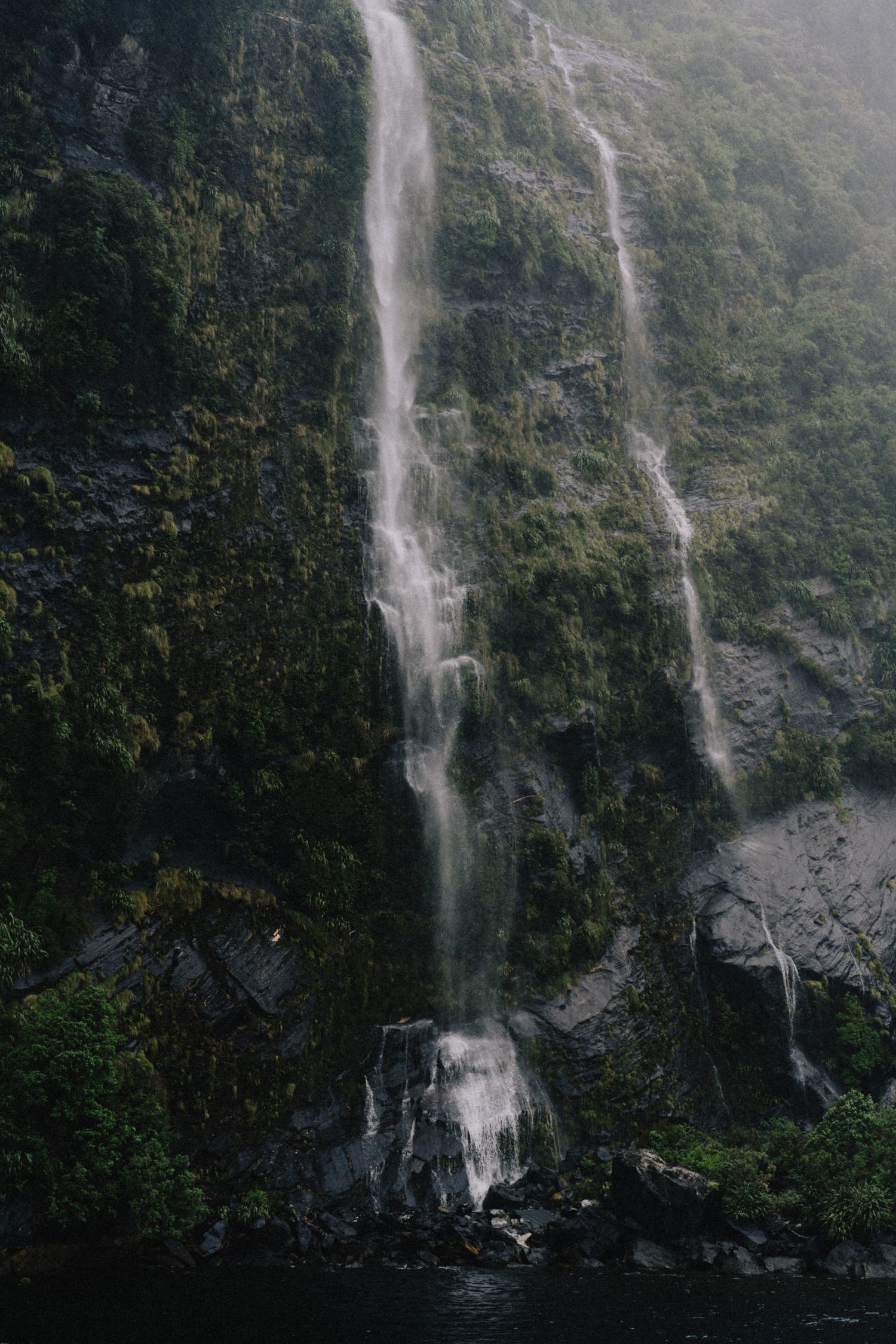 Two dramatic waterfalls cascade down a steep moss-covered cliff face in a misty forest landscape.