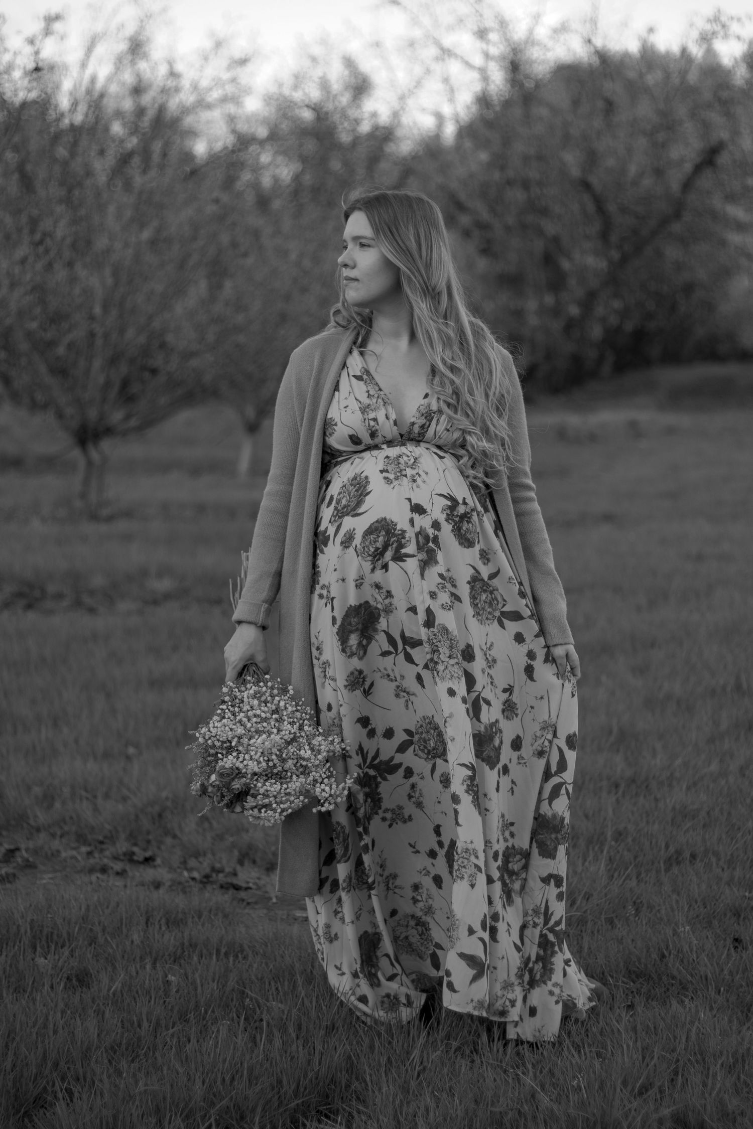 Expectant mother in floral dress holding wildflowers in orchard at sunset.