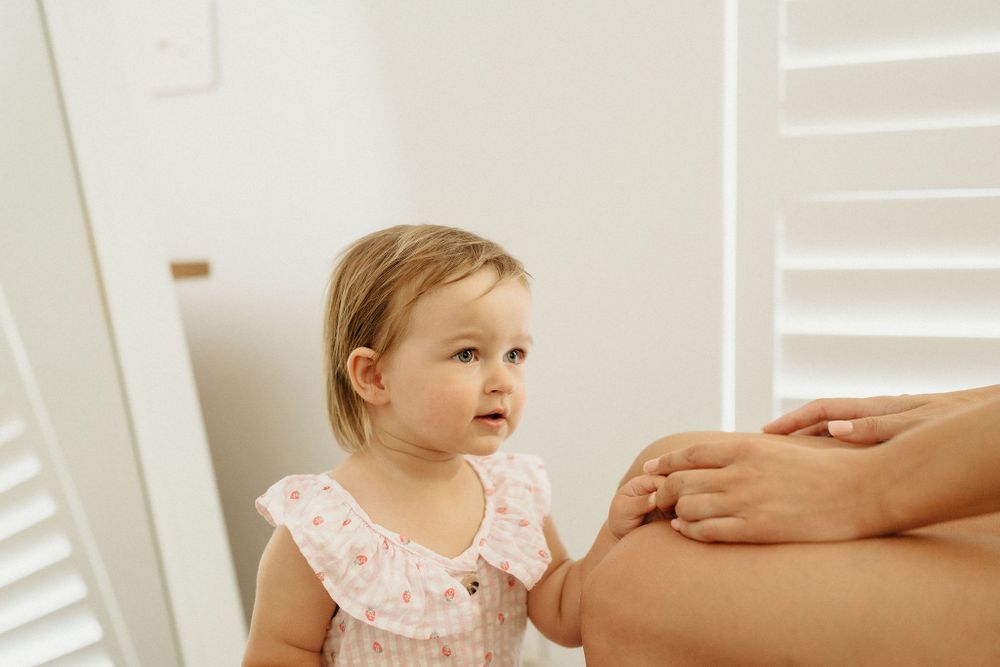 A toddler in a pink ruffled dress looks up curiously at an adult's pregnant belly.