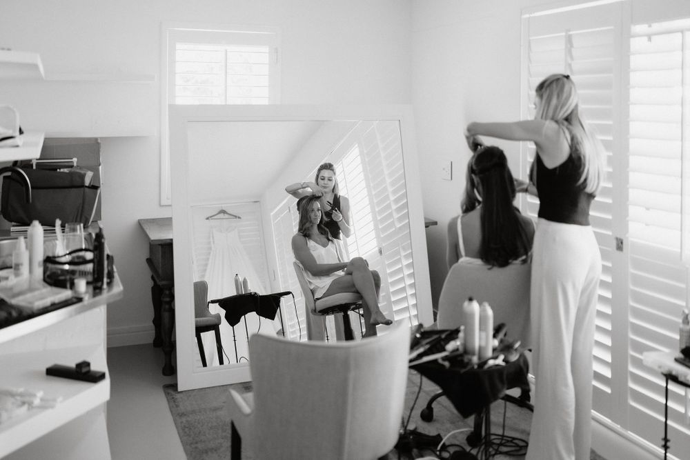 Black and white photo of preparation in a bright room with shuttered windows and styling chairs.