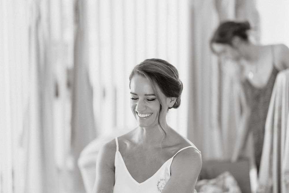 Black and white photograph showing a bride getting ready with a joyful smile while someone helps in the background.