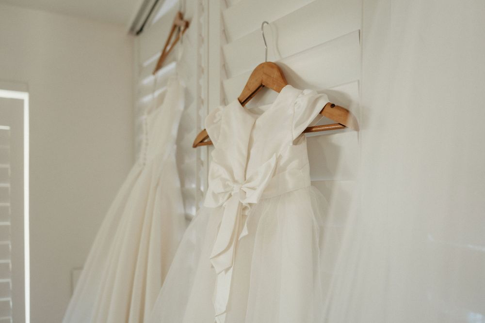 Two elegant white wedding dresses hanging on wooden hangers against a light wall.