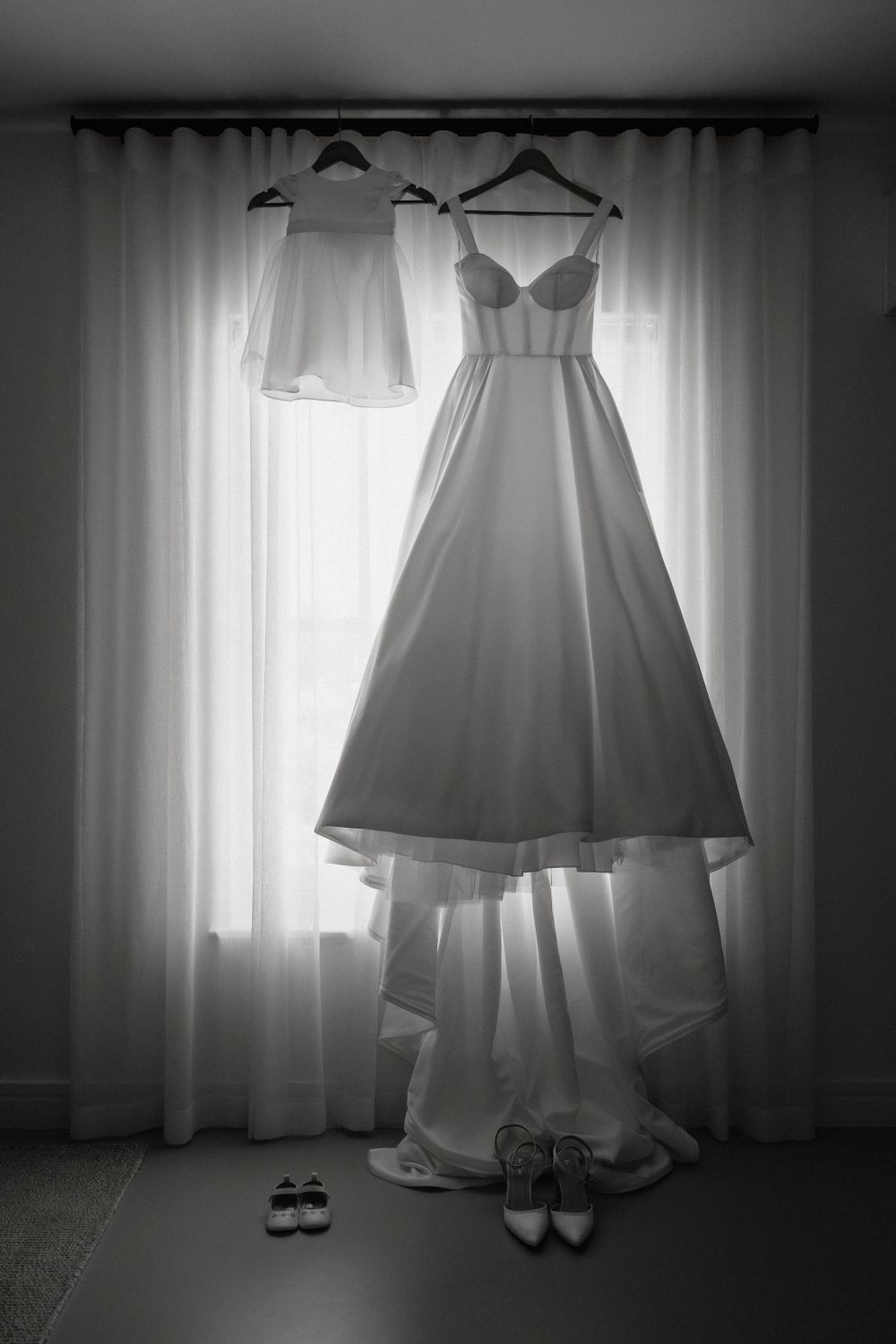 A wedding dress and smaller white dress hang from hangers against a window, with bridal shoes beneath on the floor.