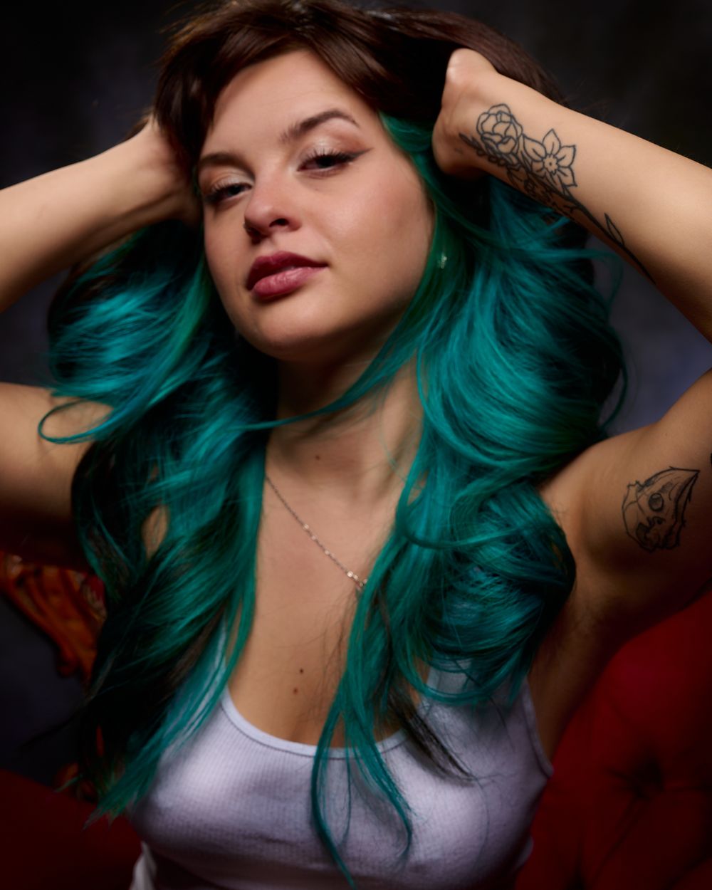 Fashion model with vibrant teal hair poses expressively in a white tank top against a dark background.