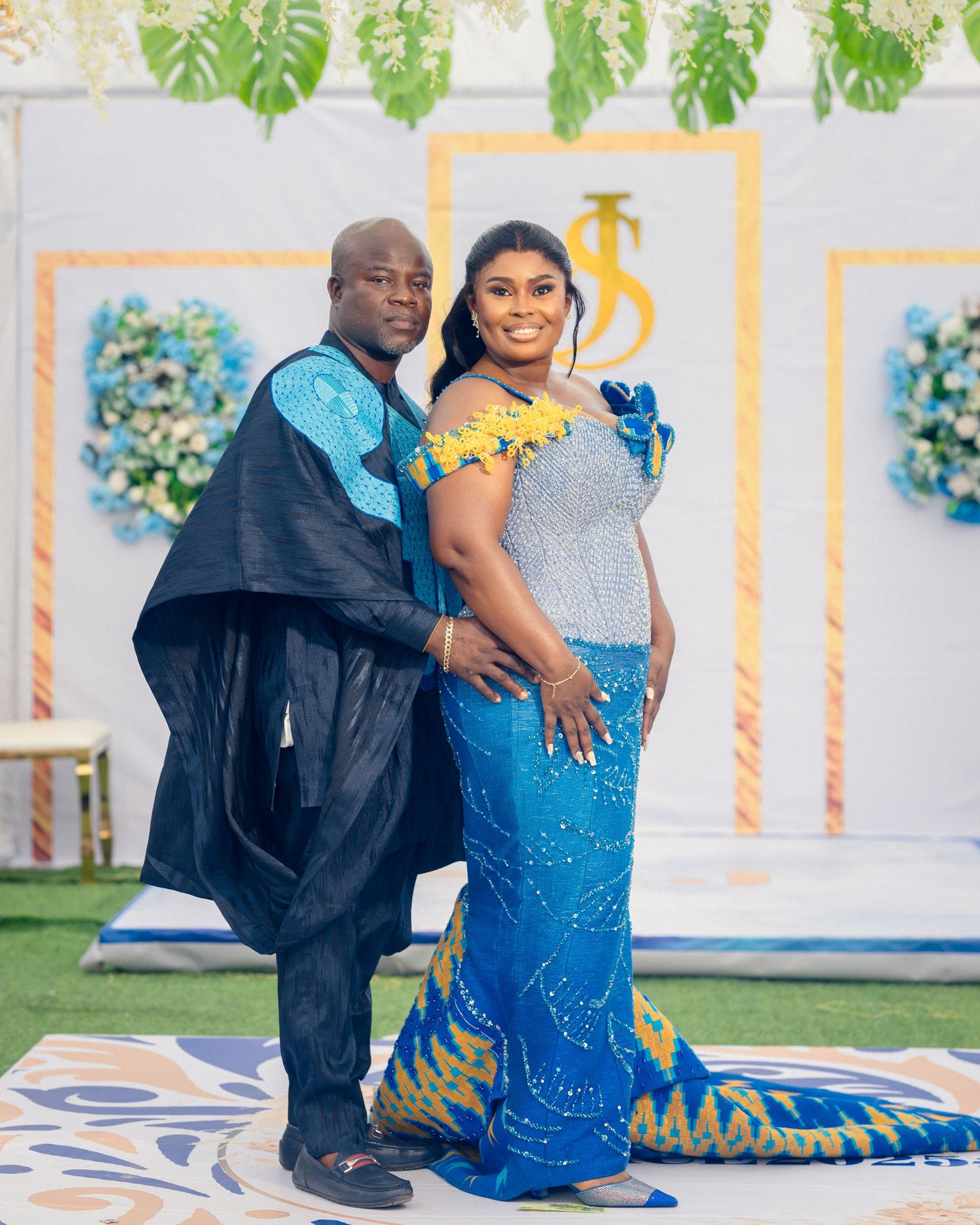 Couple poses in coordinating blue outfits against a decorated wedding backdrop with tropical leaves.
