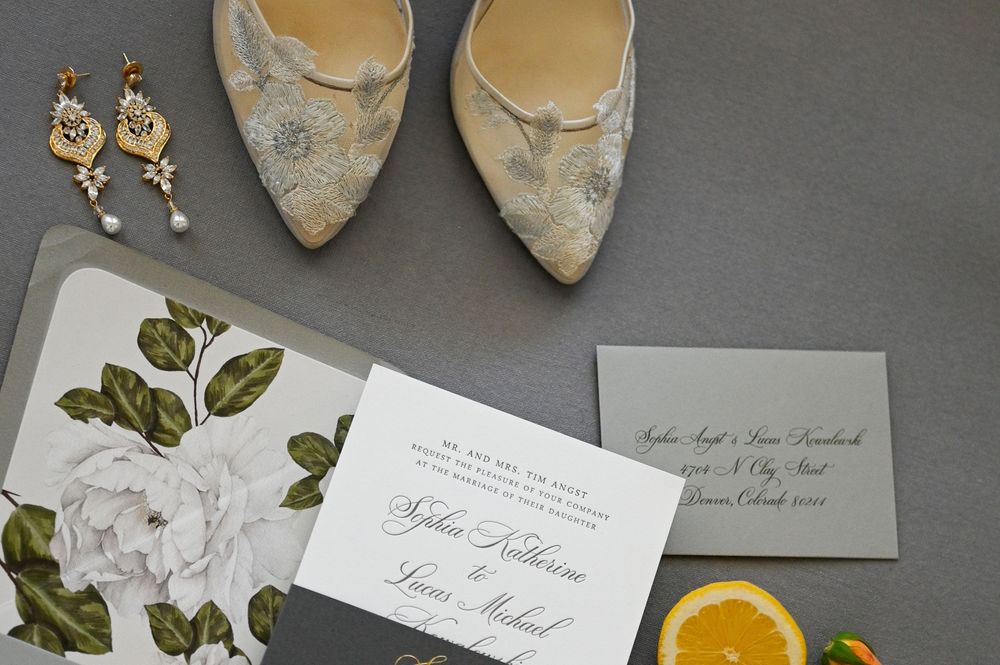 Wedding invitation suite with beige lace shoes, floral stationery, gold earrings and a citrus slice on gray surface.