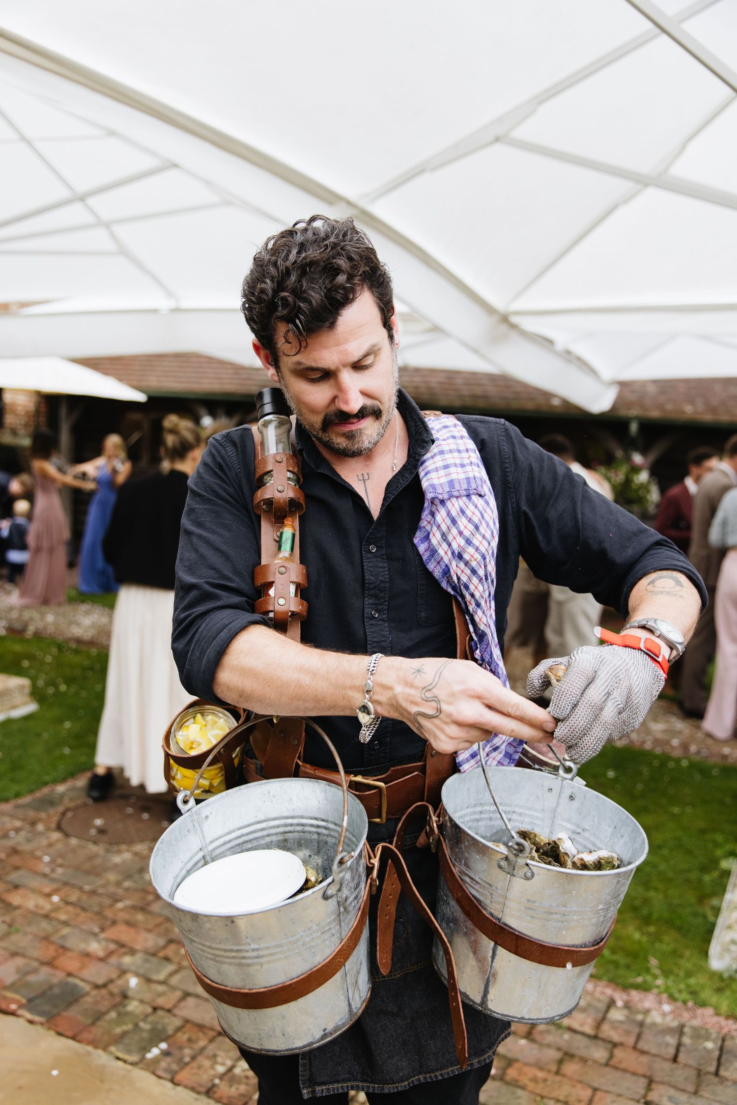 Server wearing black shirt and suspenders carries metal buckets while serving food at an outdoor event with tent canopy.
