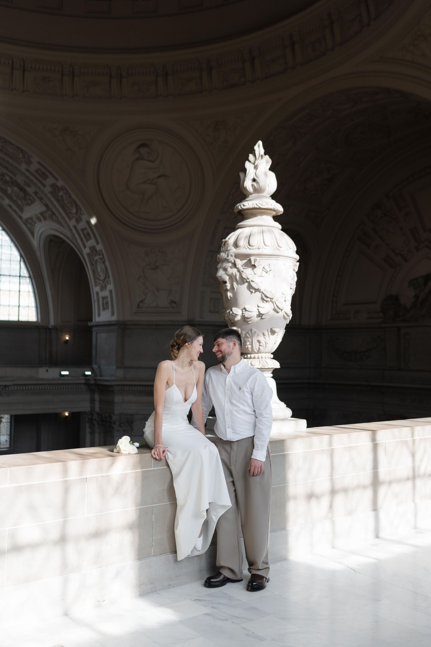 A couple poses together on a marble balcony inside an ornate classical building with decorative architectural details.