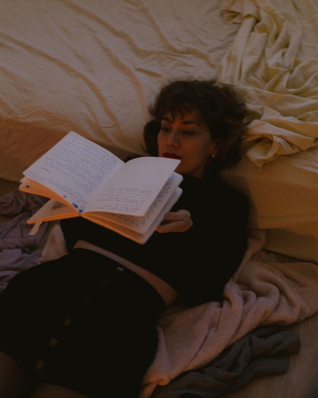 A young woman lying in bed reading a book by warm lamplight in a cozy bedroom setting.
