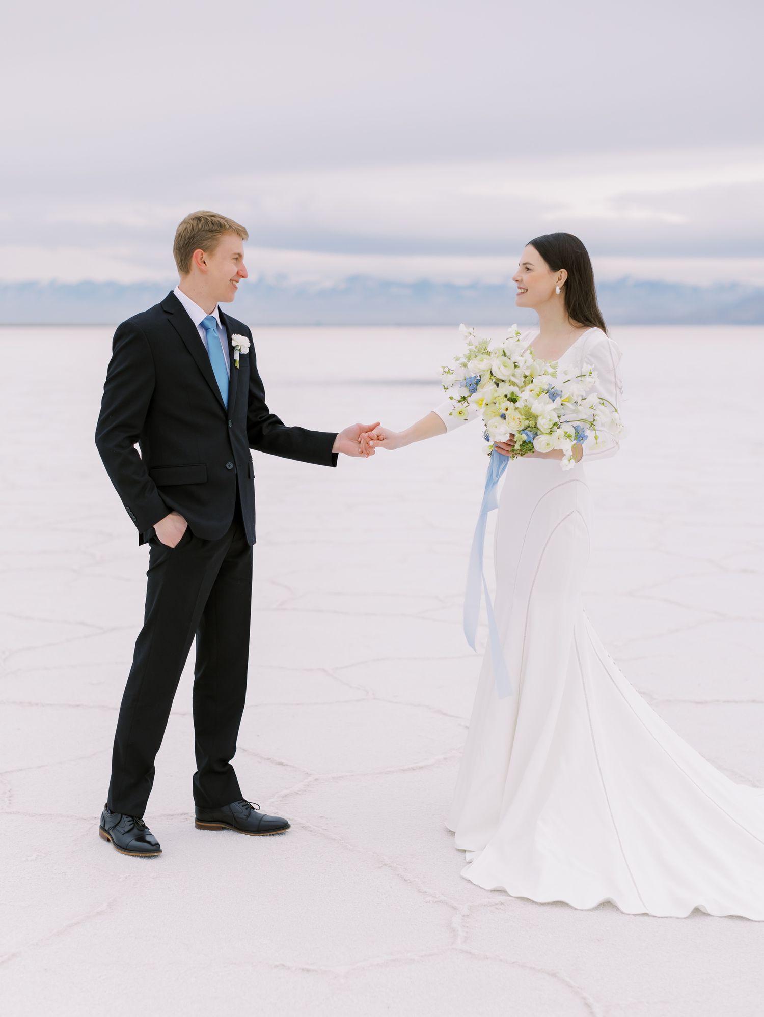 A newlywed couple stands together on a salt flat landscape under cloudy skies with mountains in the distance.