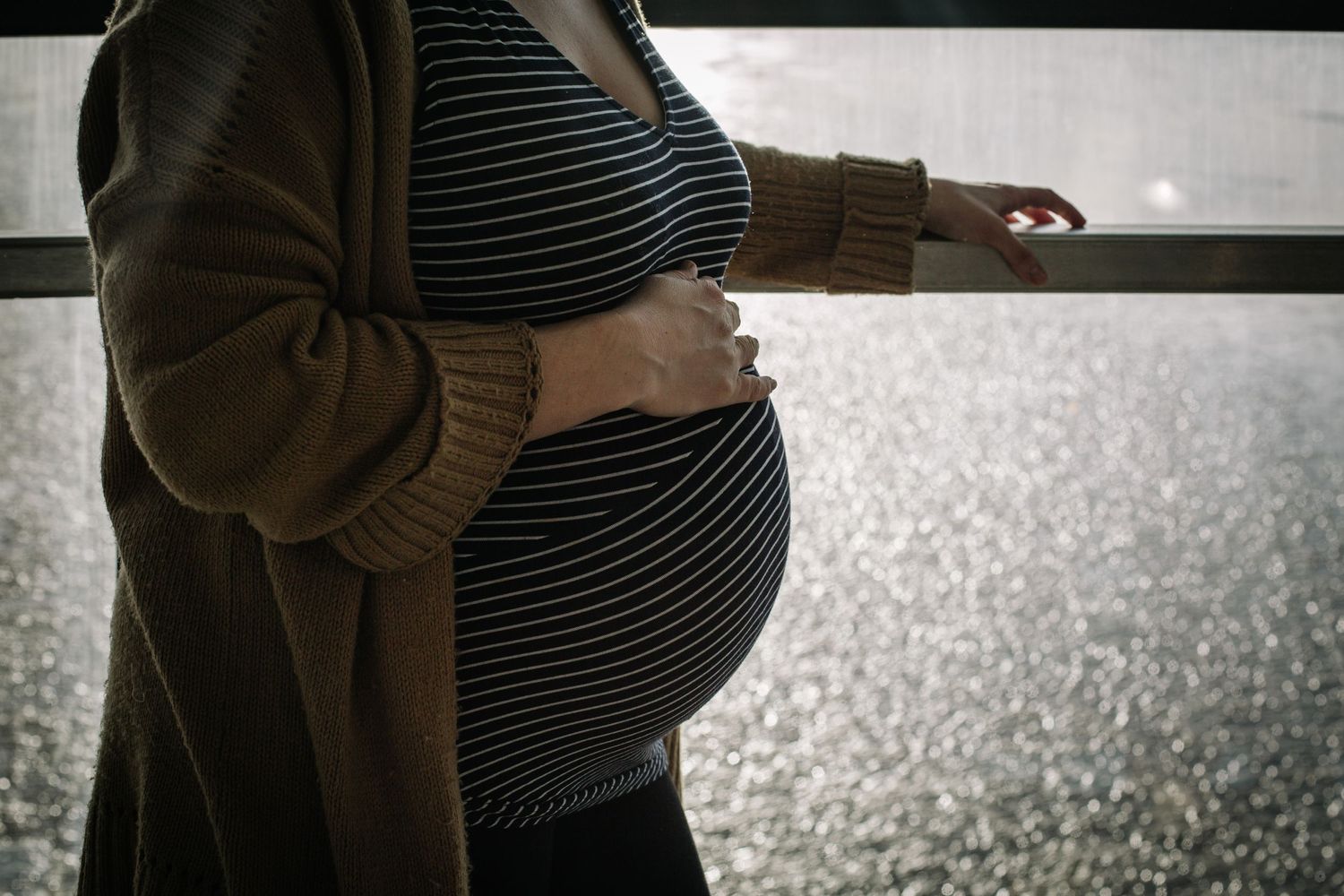 A pregnant person wearing a brown cardigan and striped dress stands by a window, holding their baby bump.
