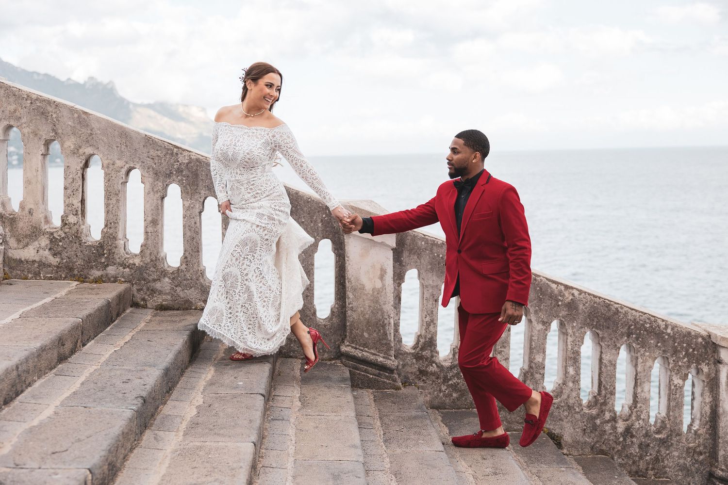 A couple in wedding attire poses on stone steps by the ocean, with the bride in white and groom in burgundy.