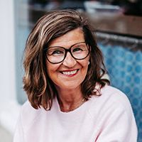 Frau mit braunen Haaren und Brille lächelt in die Kamera. Sie trägt einen rosa Pullover und sitzt vor einem blauen Hintergrund.