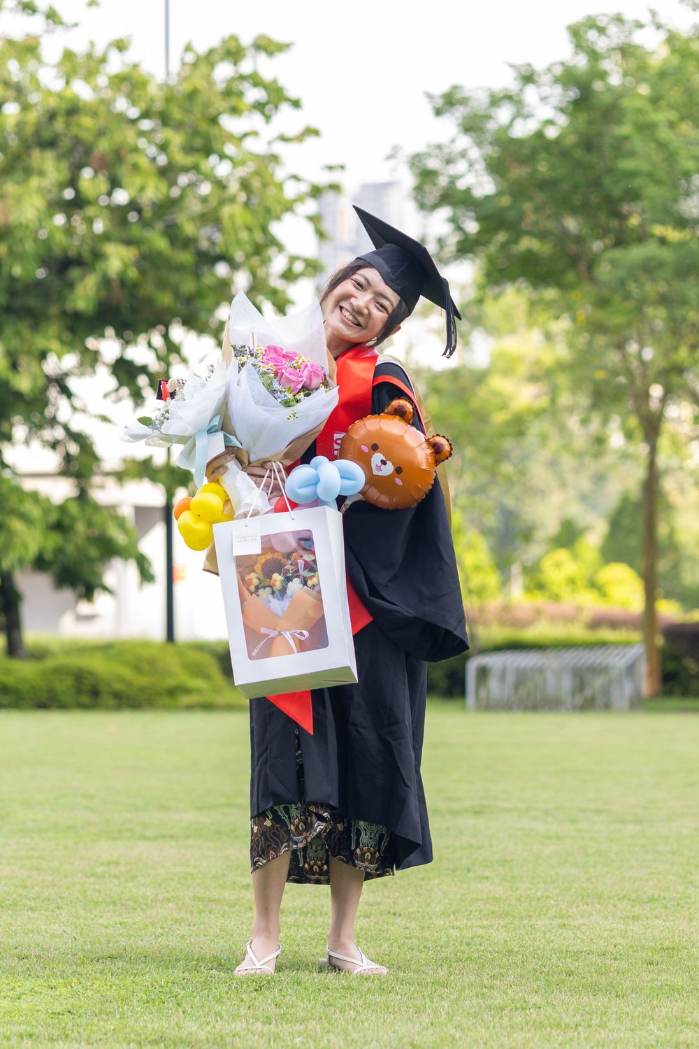Graduate in academic regalia poses with teddy bear and gifts on graduation day.