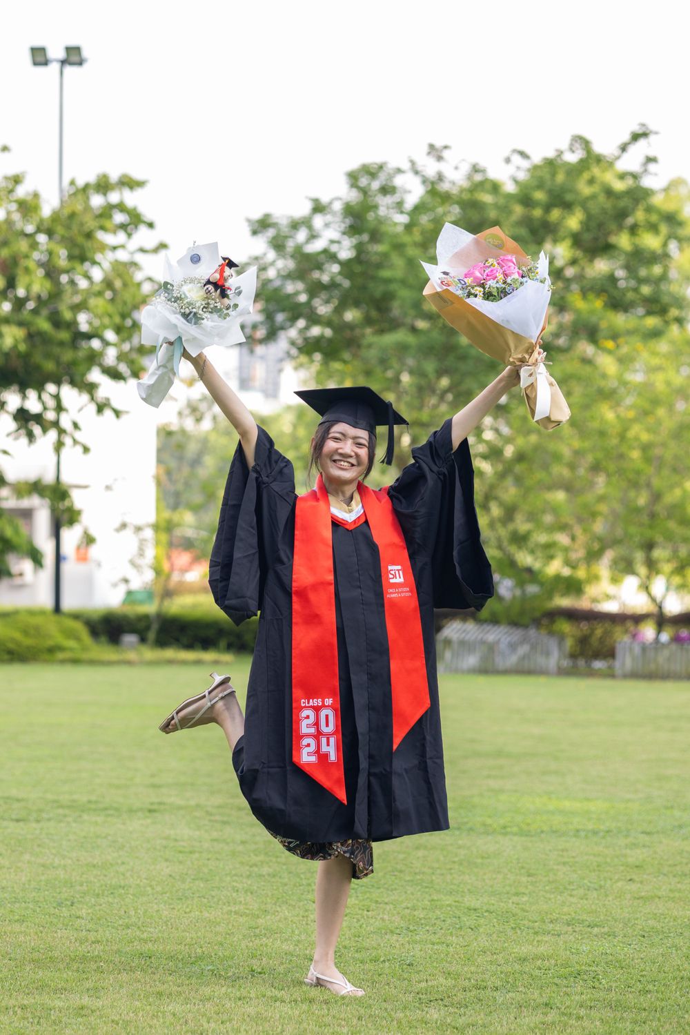 Graduate in black cap and gown with red stole celebrates on lawn while holding flower bouquets.