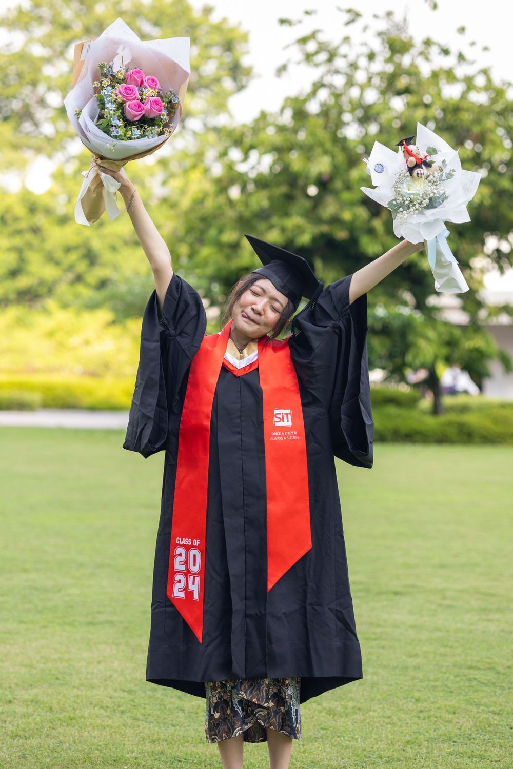 Joyful graduate holds flower bouquets aloft while wearing traditional graduation attire outdoors.