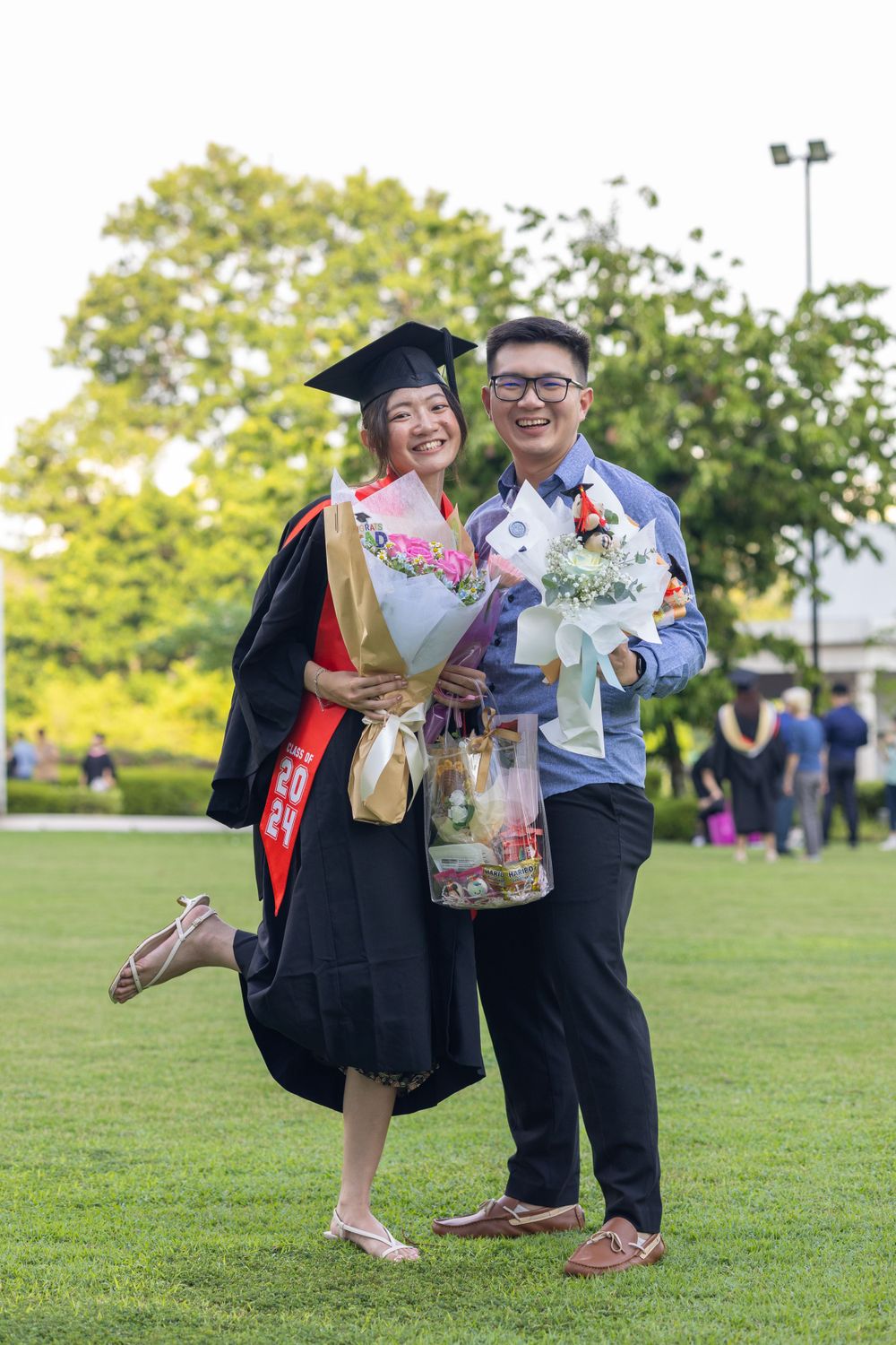 Graduation portrait of couple with flower bouquets on sunny lawn.
