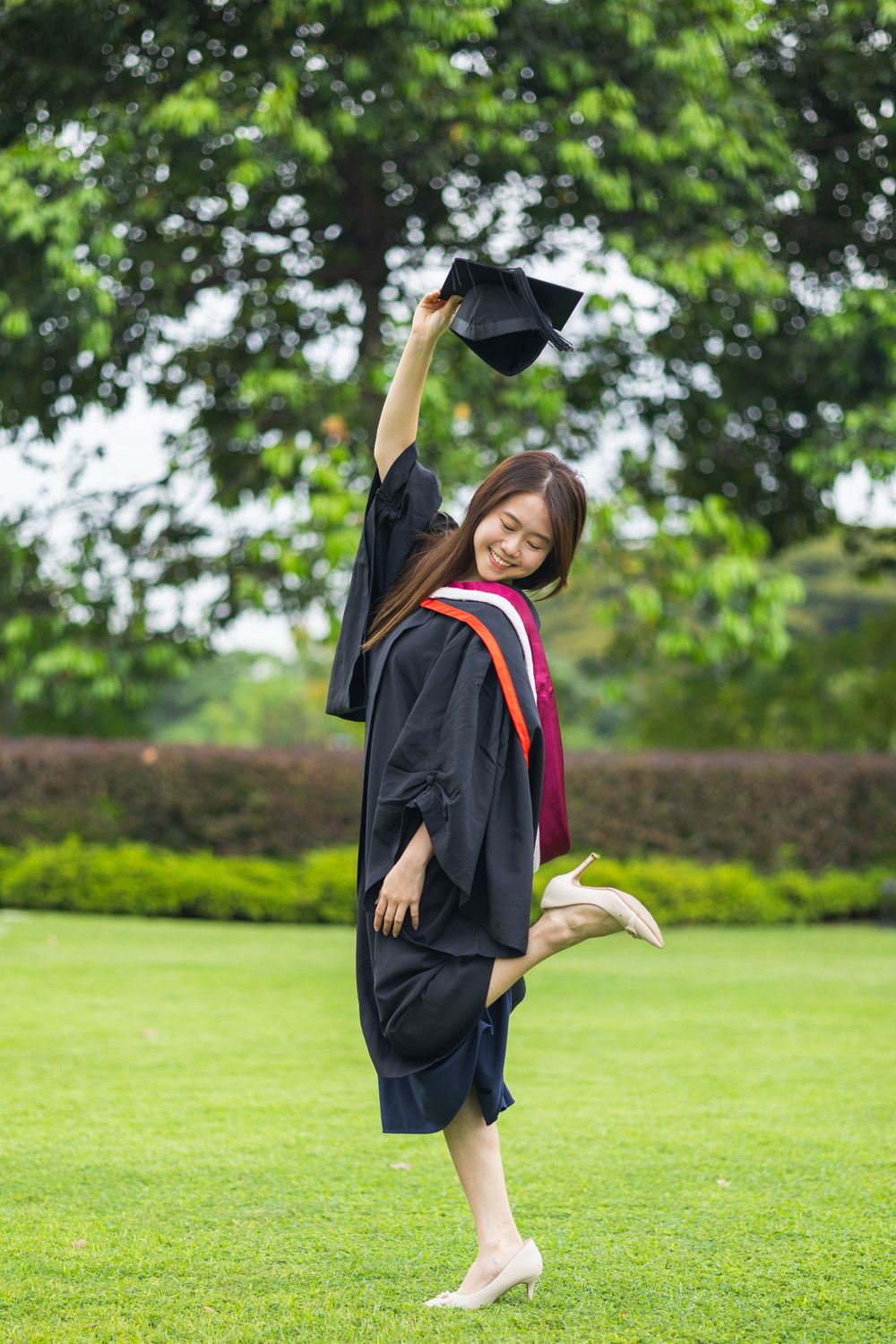 Graduate in black cap and gown strikes playful pose holding cap up on grassy lawn with trees in background.
