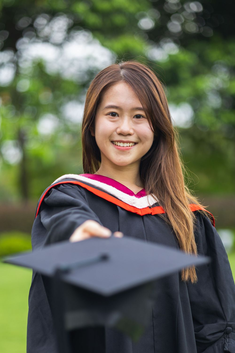A graduate in black academic regalia with red trim holds their mortarboard cap on a sunny day outdoors.