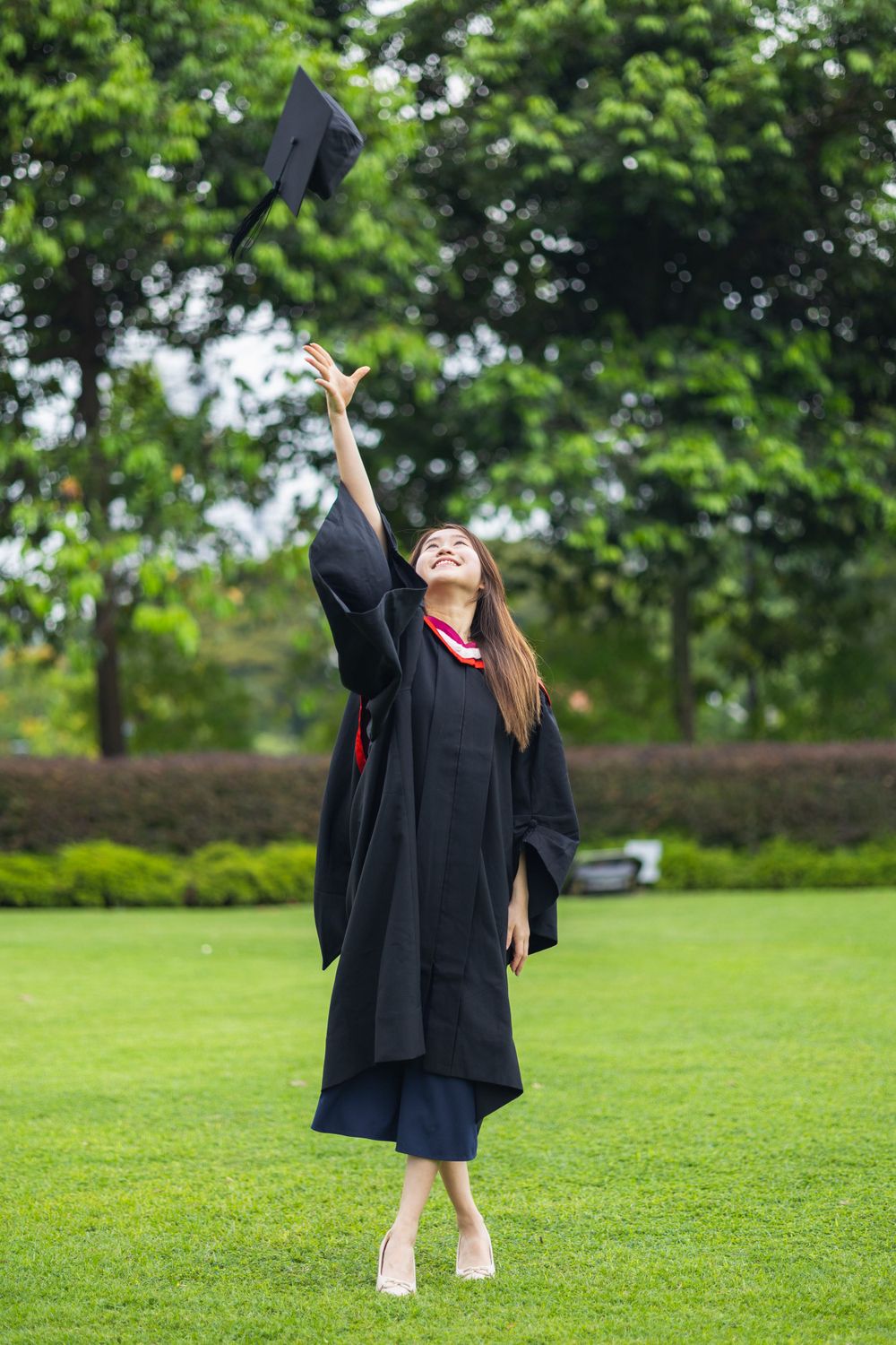 Graduate in traditional black gown throws graduation cap high in the air while standing on green lawn.