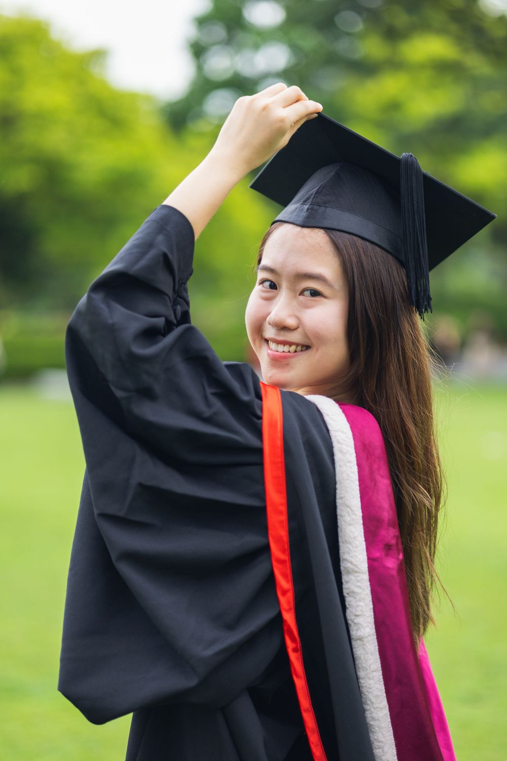 Graduate wearing academic regalia and cap adjusts mortarboard while smiling outdoors.