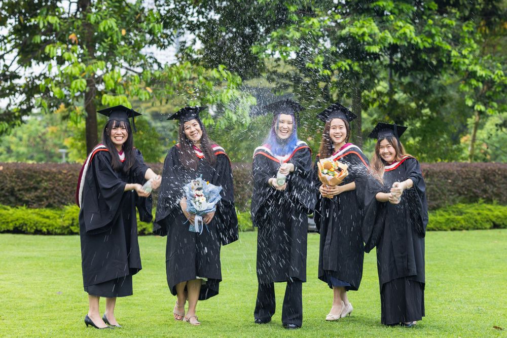 Group of graduates in academic regalia celebrate with bouquets outdoors.
