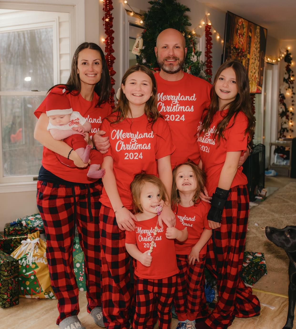 Family wearing matching red shirts and buffalo plaid pajama pants poses by Christmas tree with dog.