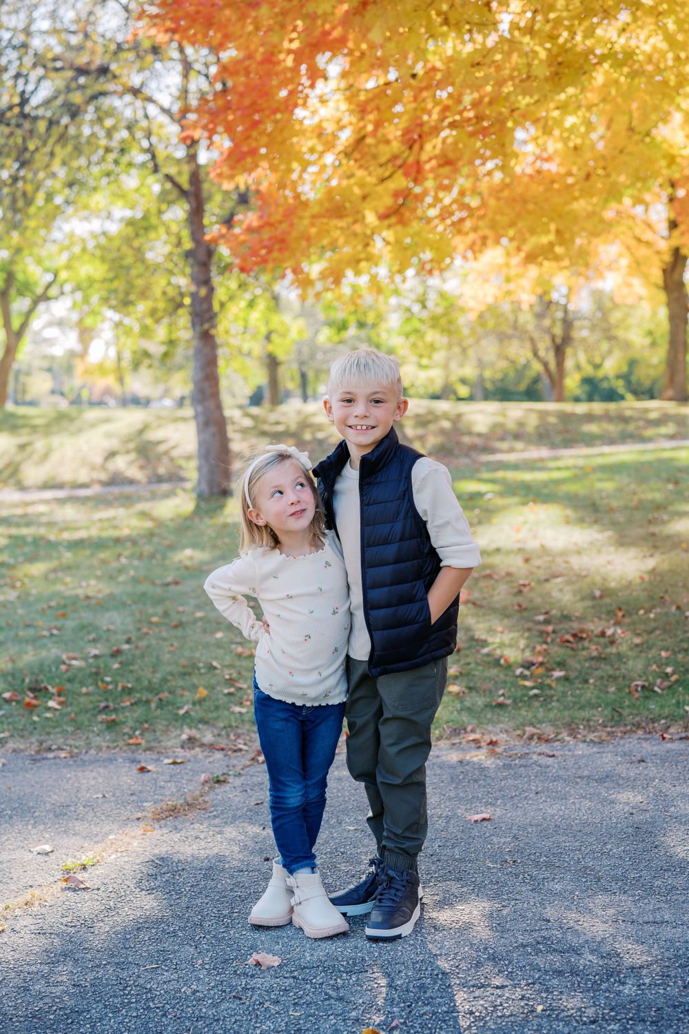 Two young children playing and smiling together on a path surrounded by autumn trees with golden foliage.