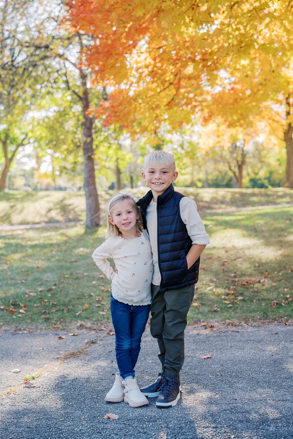 Two children standing together on a path with beautiful fall colors and trees in the background.