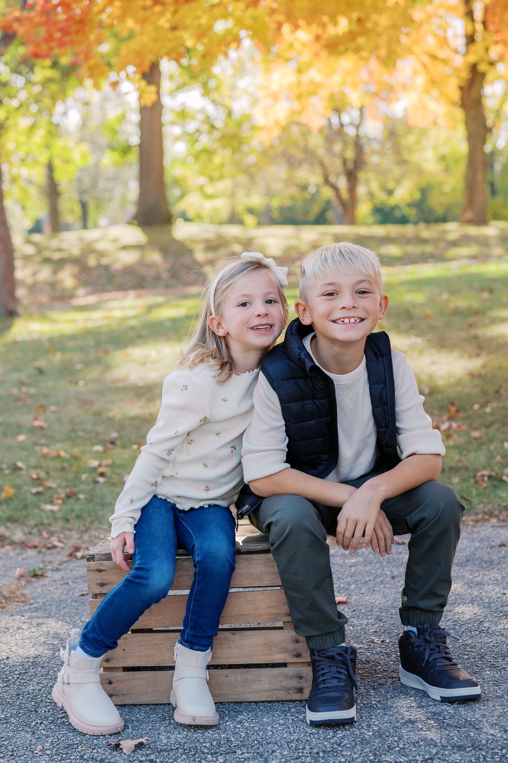 Two children sitting together on wooden crates under colorful autumn trees.