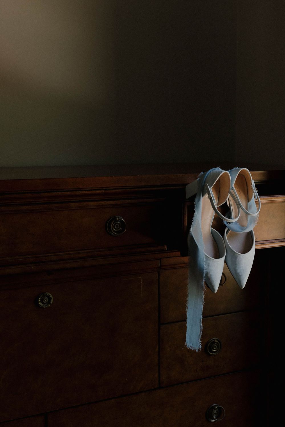 Light blue wedding shoes placed on a dark wooden dresser in dramatic lighting.