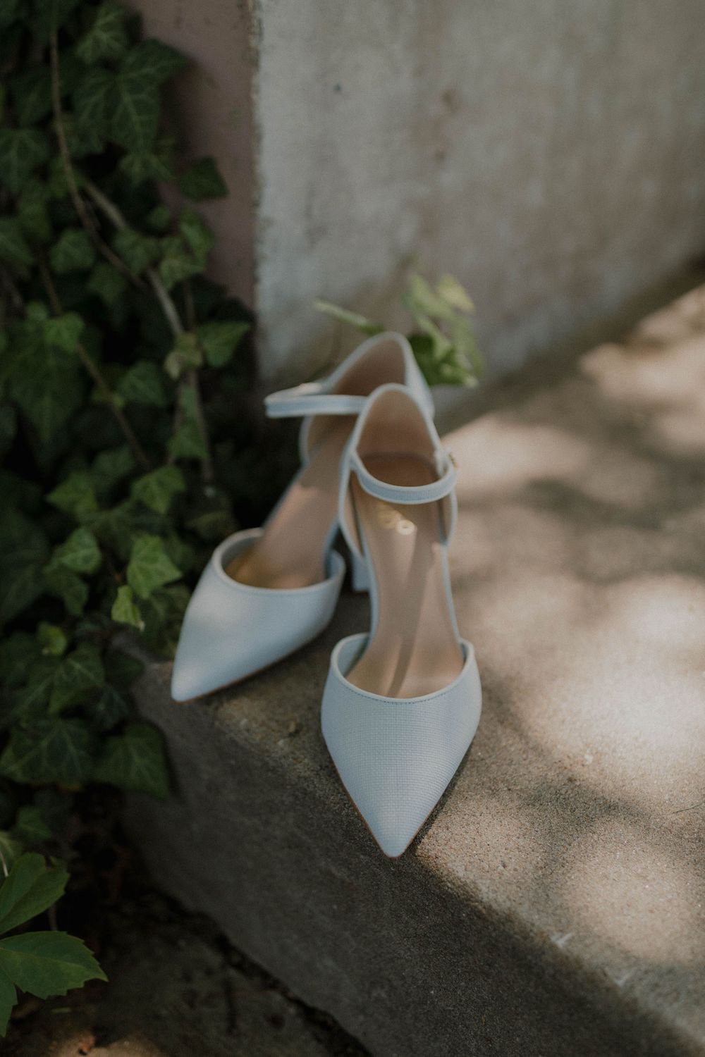 Elegant white bridal shoes with ankle straps on stone ledge in dappled sunlight.