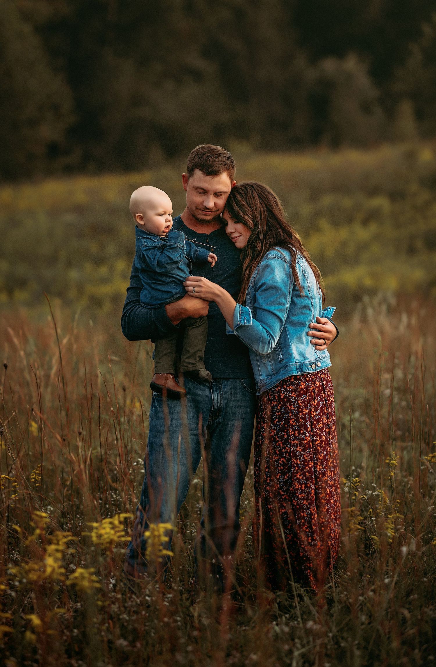 Family of three embraces in a rustic autumn meadow at sunset wearing casual denim and floral clothing.