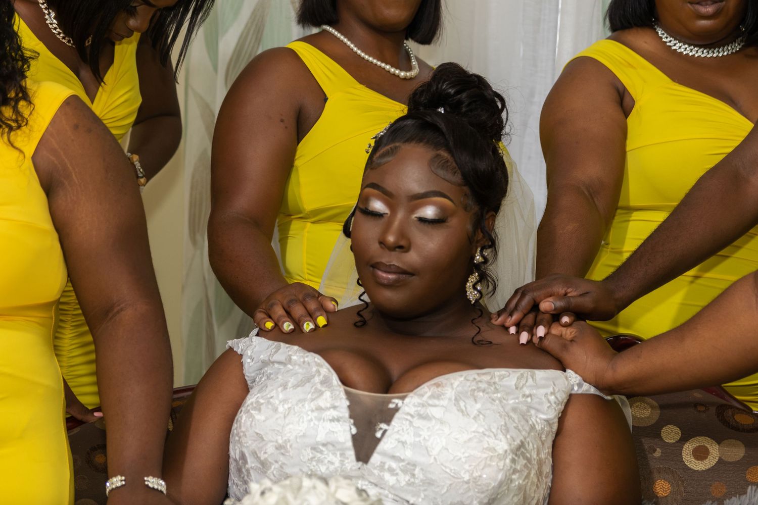 Close-up of a serene bride in a white wedding dress with intricate lace details, eyes closed, as her bridesmaids in bright yellow dresses.