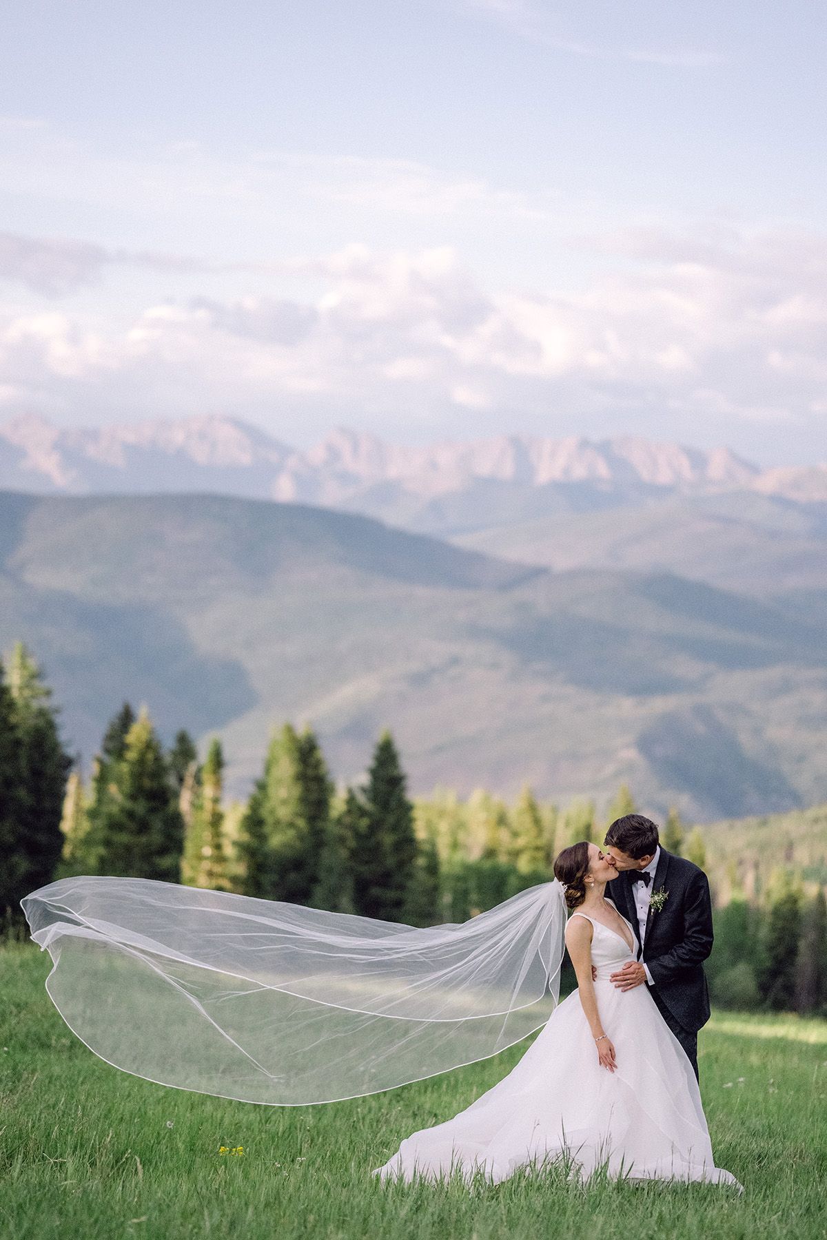 Romantic mountain wedding photo with bride's veil flowing dramatically against mountain landscape.