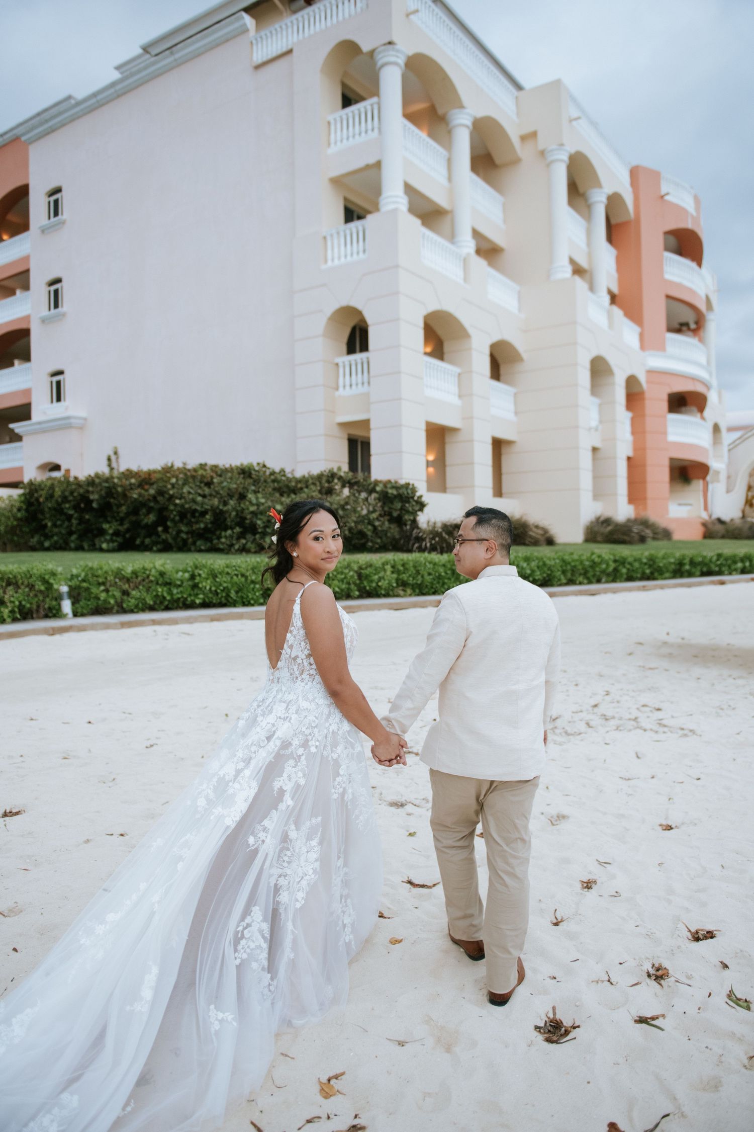 Couple in wedding attire walking hand in hand outside a pink and white Mediterranean-style resort building.