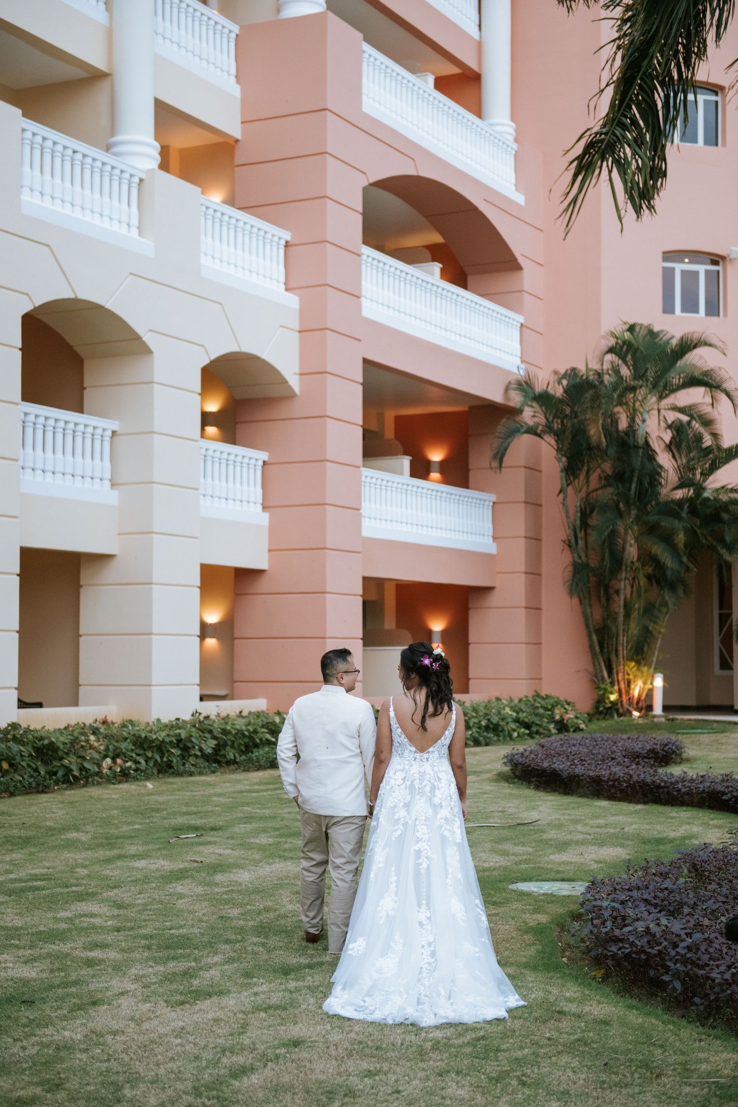 Couple walking through tropical garden path beside pink Mediterranean-style resort architecture.