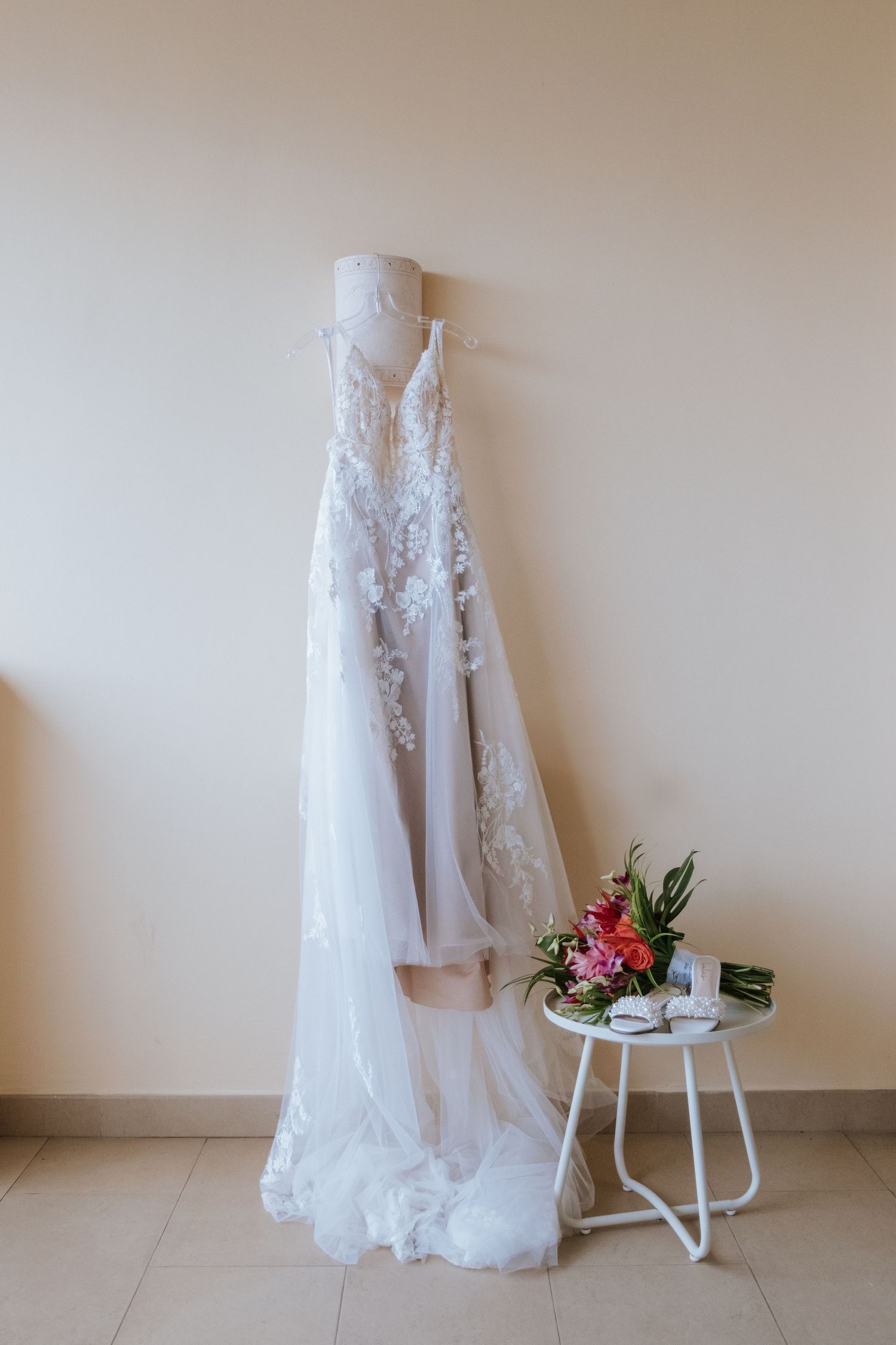 White lace wedding dress hanging on wall with small floral arrangement on side table.