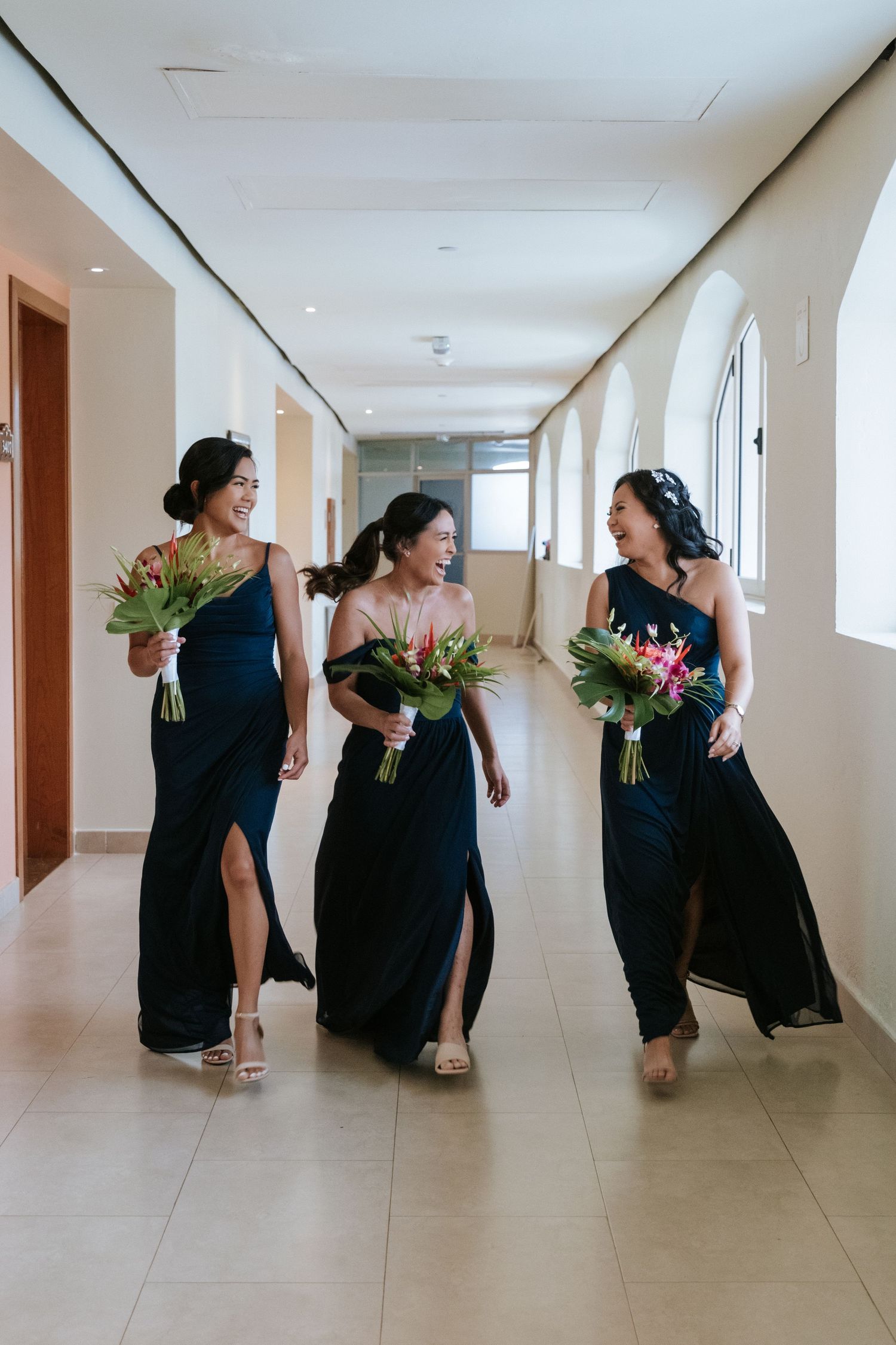 Three bridesmaids in navy dresses walking down hallway carrying floral bouquets.