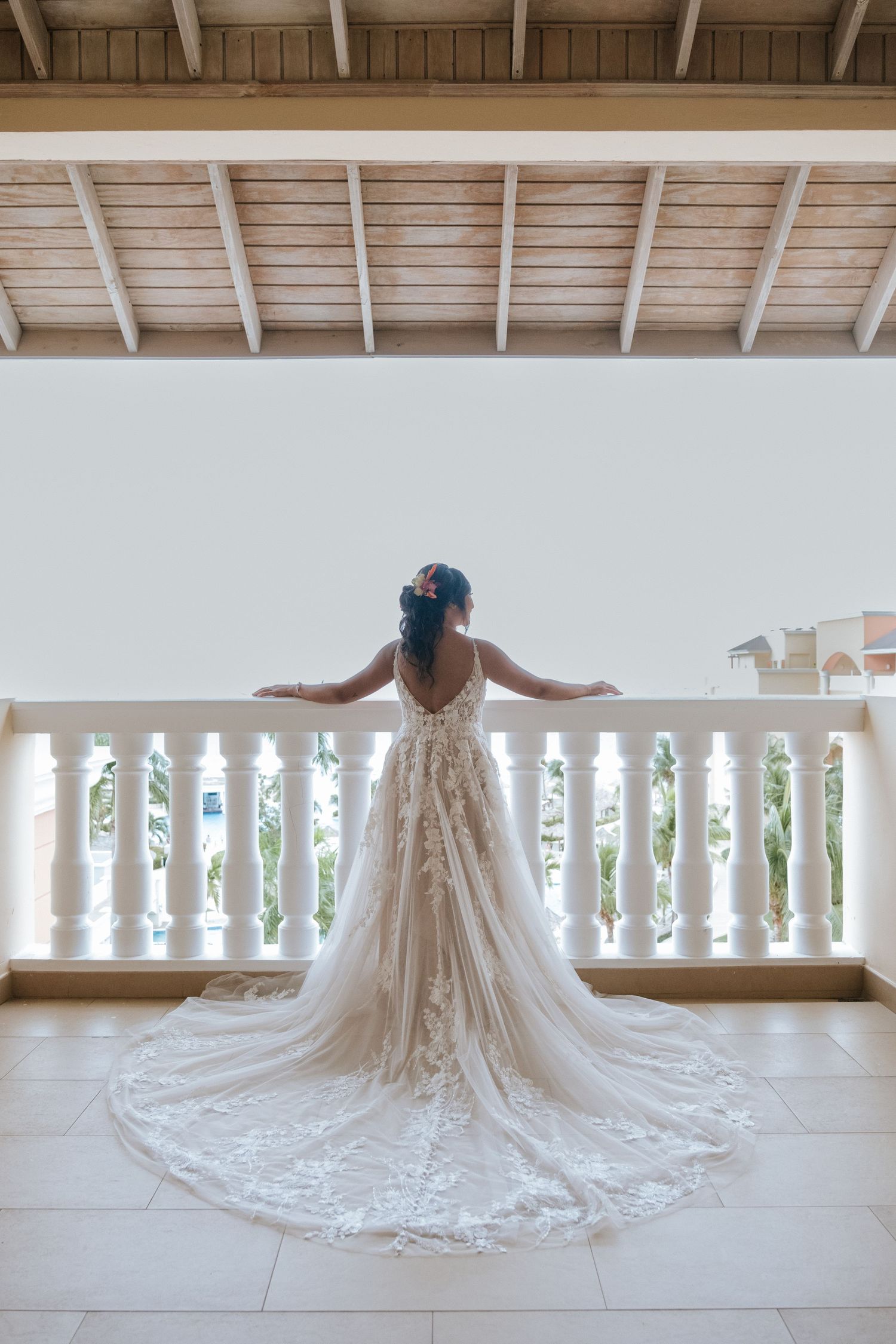 A bride in a lace wedding dress with long train stands at a white balcony railing overlooking a resort.