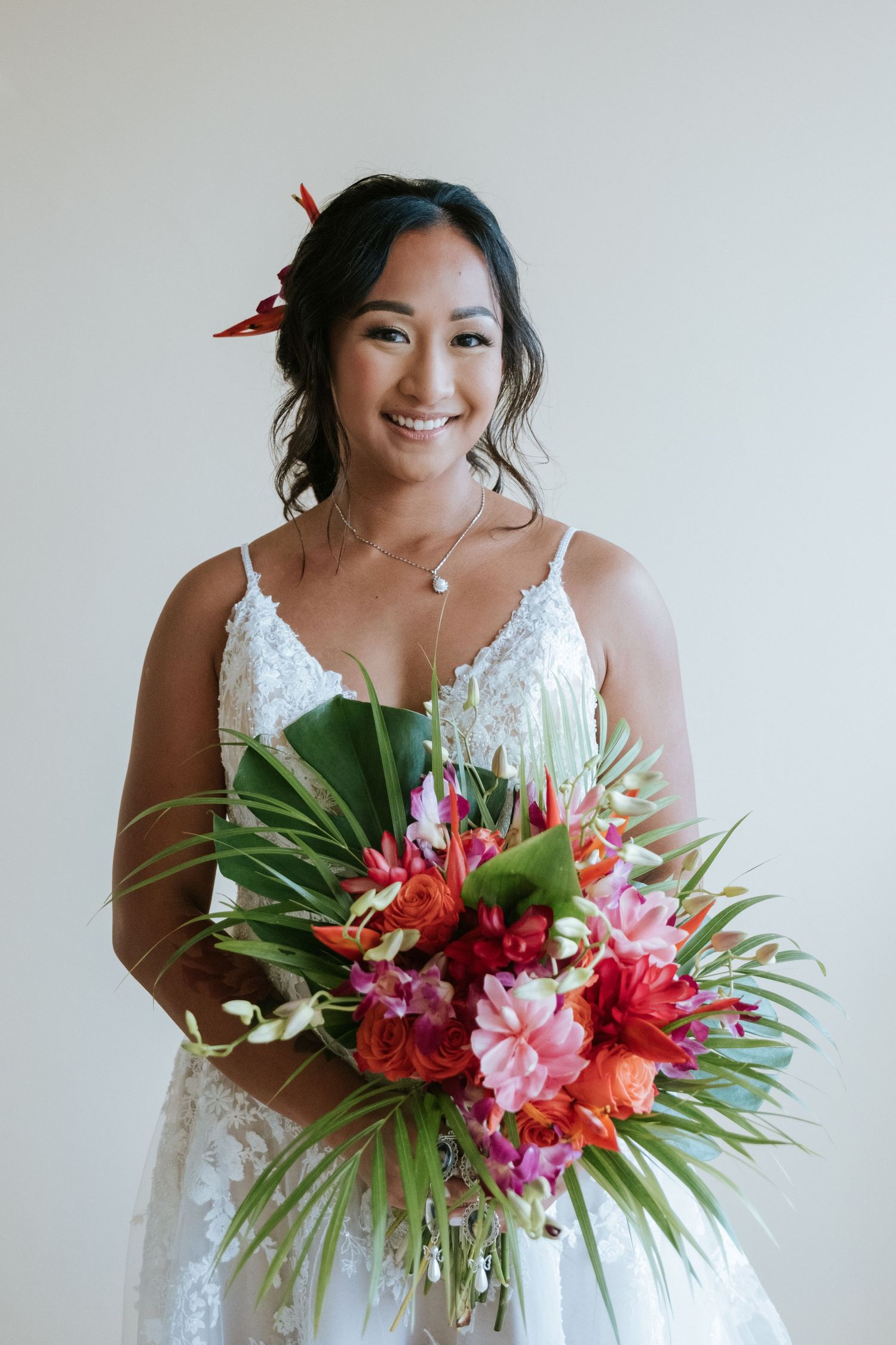 A bride holding a vibrant tropical bouquet of red and pink flowers with green palm leaves.