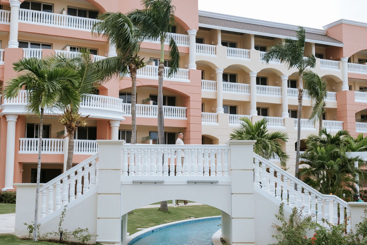 A pink resort building with white balconies and a decorative bridge over a swimming pool.