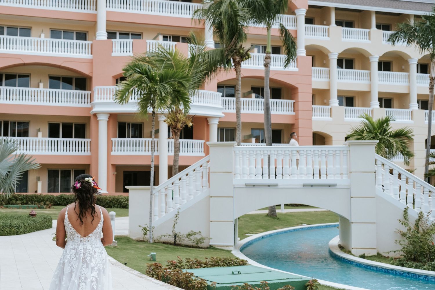 A person in a white dress walking near a decorative bridge at a pink resort building.
