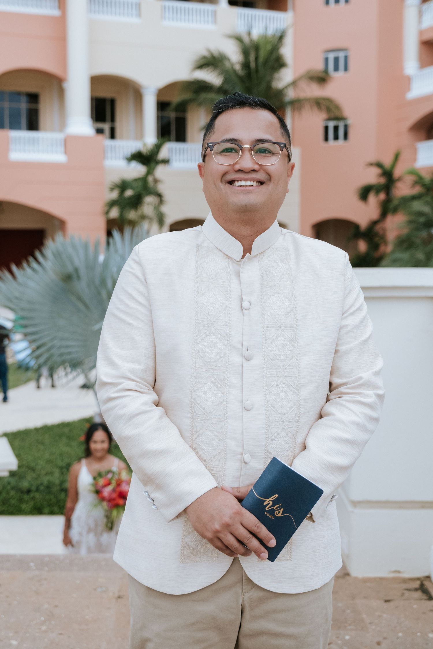 A person in white formal attire holding a blue book at a tropical resort.