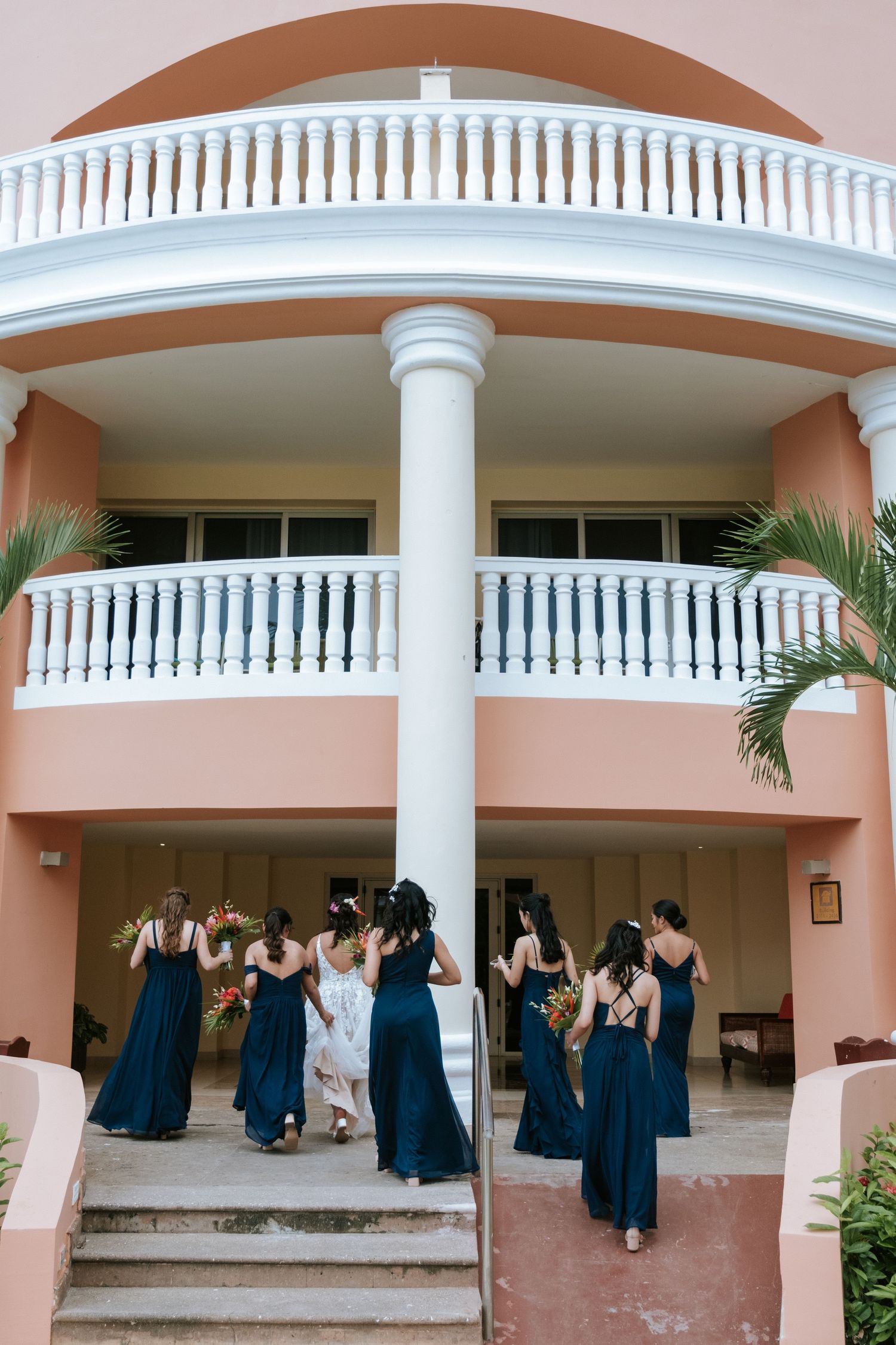 A group in navy blue dresses gather on the steps of a pink Mediterranean-style building with white columns and balconies.
