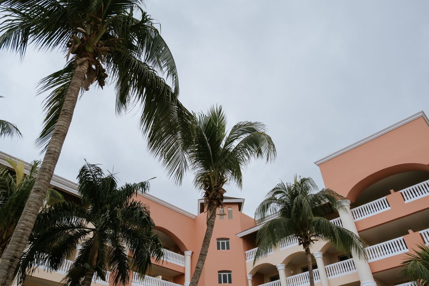 Pink resort building with palm trees swaying against cloudy sky.