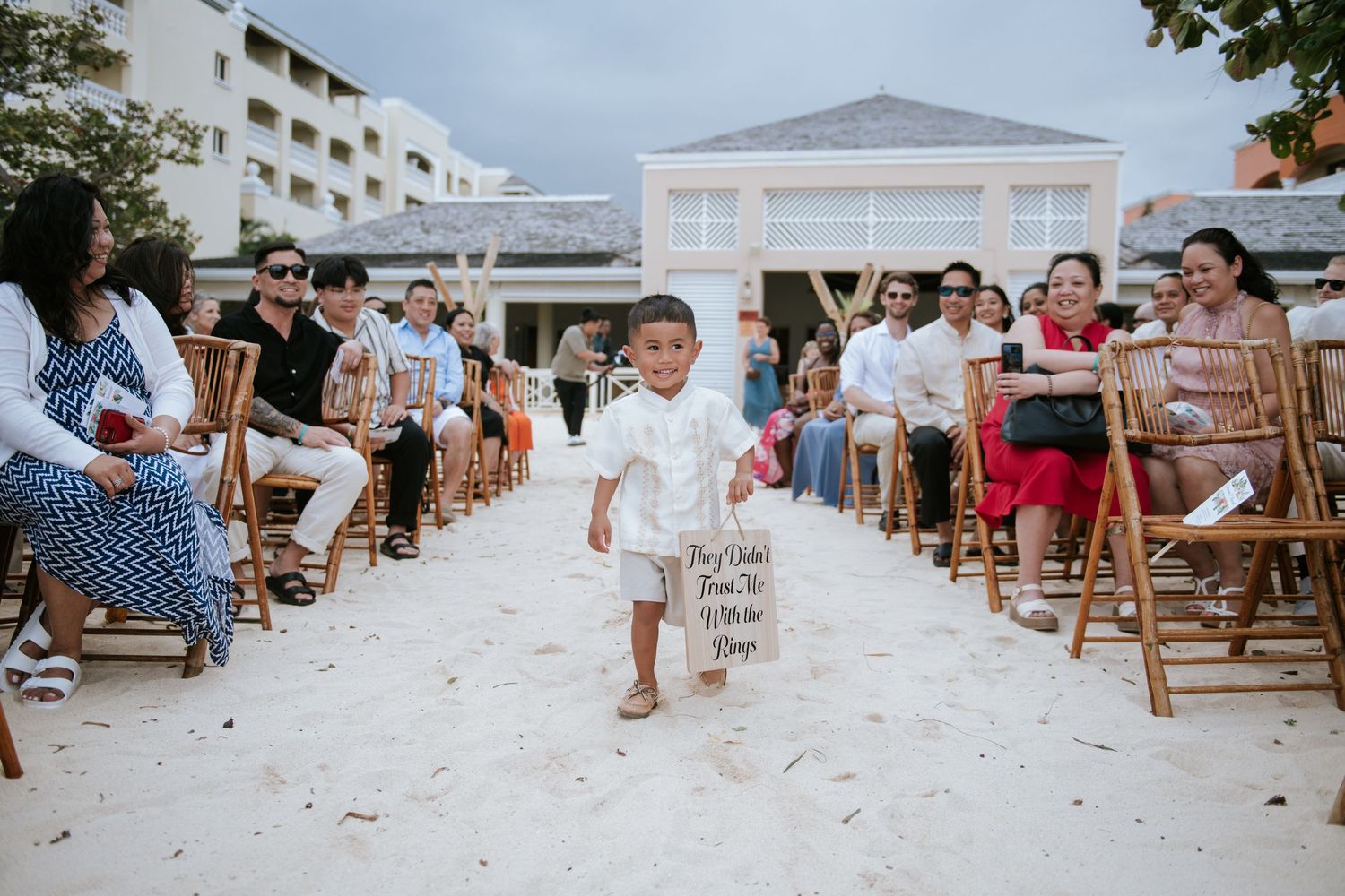 A beach wedding ceremony with wooden chairs lined up on white sand and guests watching.